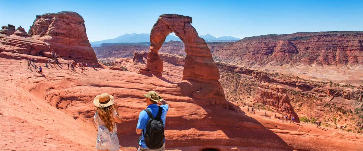 Couple on vacation hiking trip. Man and woman standing on top of the mountain looking at beautiful view. Delicate Arch, Moab, Utah, Arches National Park.